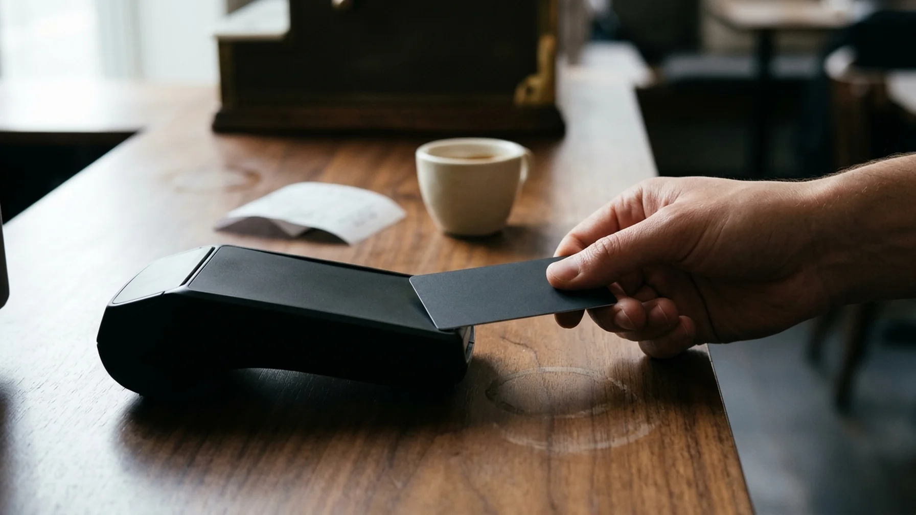 Close-up of a customer's hand tapping a contactless bank card against a payment terminal on a café counter, with a paper receipt and a ceramic cup nearby